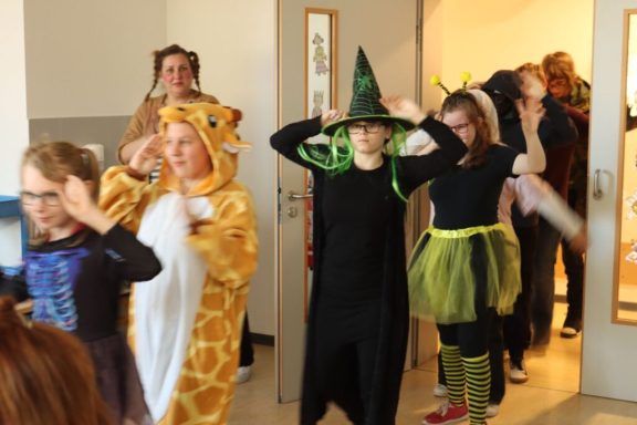 Kinder in Kostümen, darunter eine Giraffe und eine Hexe, ziehen durch einen Gang.