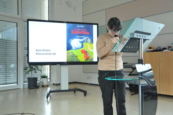 Person spricht an einem Pult, präsentiert ein Buch über Pferde. Präsentation im Hintergrund.