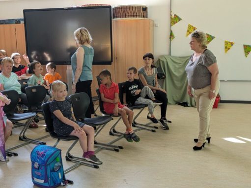 Gruppensitzung mit Kindern, Lehrerin steht vorne, viele Stühle, fröhliche Atmosphäre.
