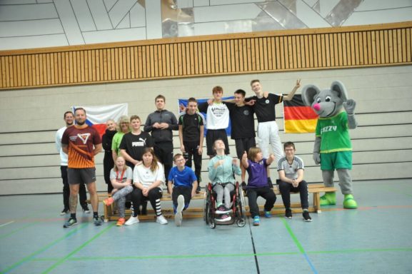 Gruppenfoto von Sportlern und einem Maskottchen in einer Halle, mit Flaggen im Hintergrund.