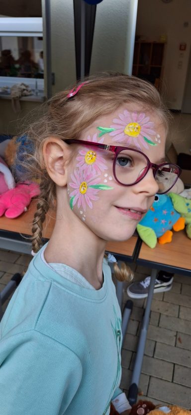 Mädchen mit buntem Gesichtsmalerei und Brille, lächelnd zur Seite blickend.