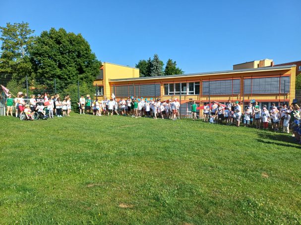 Gruppenszene mit vielen Menschen im Freien vor einem gelben Gebäude und blauen Himmel.