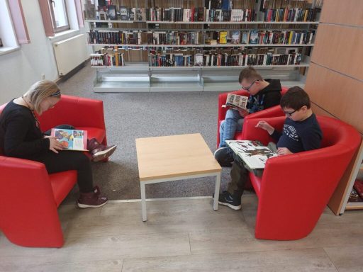 Drei Personen sitzen in roten Sesseln und lesen Hefte, Tisch in der Mitte.