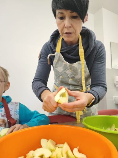 Frau schält einen Apfel, während ein Kind daneben zuschaut. Schalen liegen in einer Schüssel.