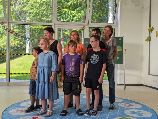 Eine Gruppe von Kindern und Erwachsenen steht lächelnd auf einem bunten Teppich.