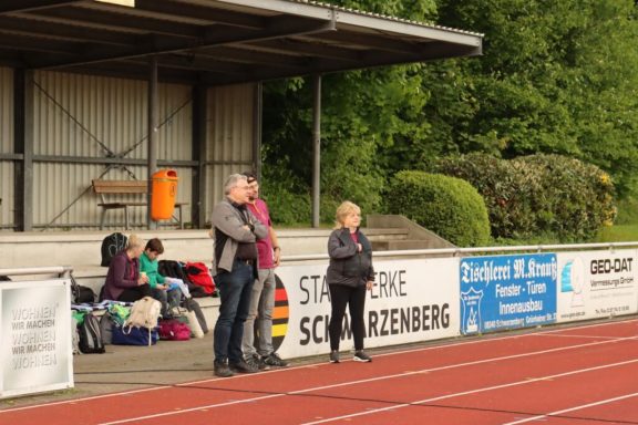 Zwei Personen stehen am Rand eines Sportplatzes, im Hintergrund sind Zuschauer und eine Tribüne sichtbar.