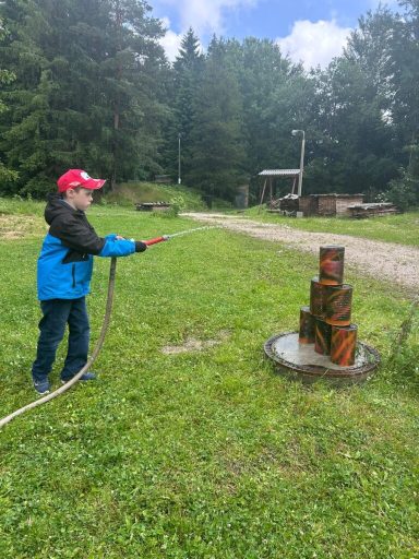Ein Junge in blauer Jacke zielt mit einem Schlauch auf einen Stapel bunter Fässer.