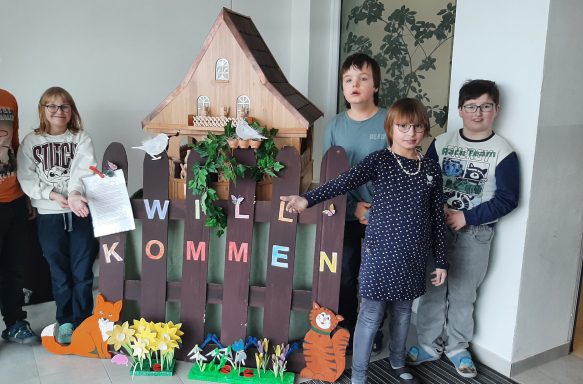 Gruppe von Kindern vor einer bunten, dekorierten Zaun aus Pappe mit dem Schriftzug „Willkommen“.