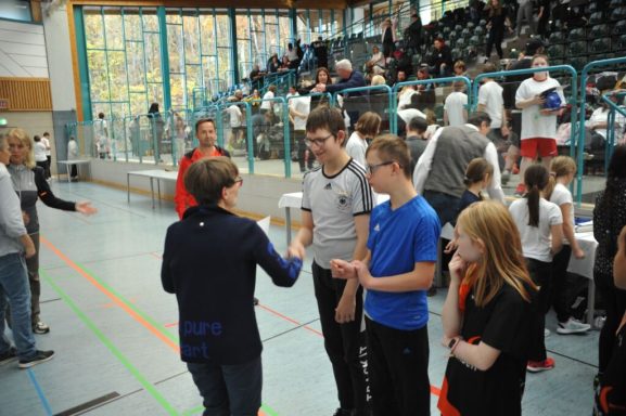 Gruppengespräch in einer Sporthalle mit Teilnehmern und Zuschauern im Hintergrund.