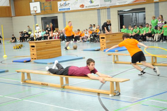 Zwei Sportler absolvieren einen Hindernisparcours in einer Halle, umgeben von Zuschauern.
