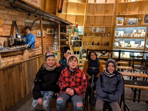 Sechs Kinder sitzen in einer gemütlichen, rustikalen Umgebung mit Holzregalen.