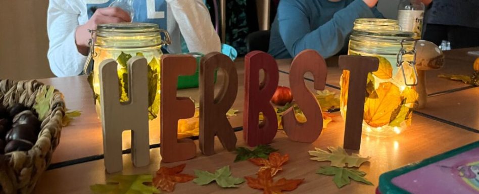 Bunte Buchstaben bilden das Wort "HERBST" vor einer herbstlichen Dekoration.