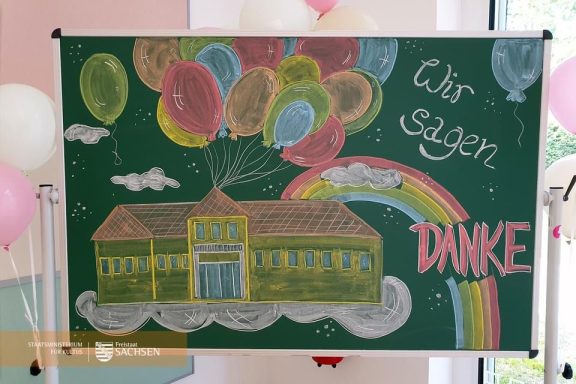 Bunte Ballons und eine farbenfrohe Zeichnung eines Hauses mit dem Text „Wir sagen Danke“.