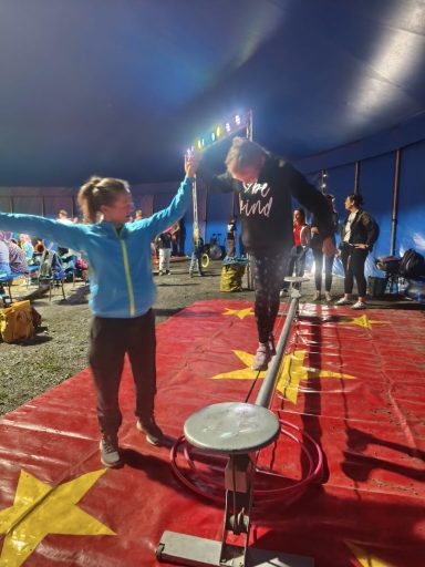 Zwei Personen im Zirkuszelt, eine balanciert auf einem Seil, die andere unterstützt sie.