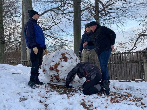 Vier Personen bauen einen Schneemann im winterlichen Schnee, umgeben von Bäumen.