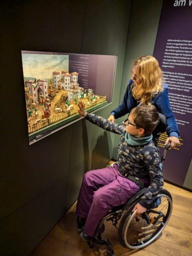 Ein Kind im Rollstuhl betrachtet mit einer Begleiterin eine Ausstellungstafel.