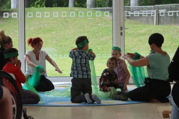 Kinder und Erwachsene spielen mit bunten Tüchern in einem hellen Raum.