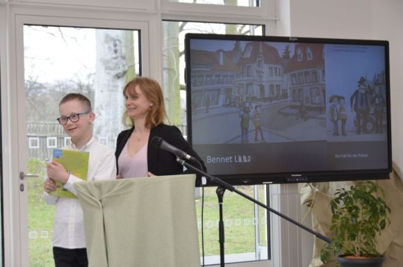 Ein Junge mit Notizen steht neben einer Frau an einem Rednerpult vor einem Bildschirm.