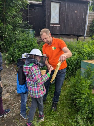 Ein Mann zeigt Kindern einen Bienenstock auf einem grünen Gelände.
