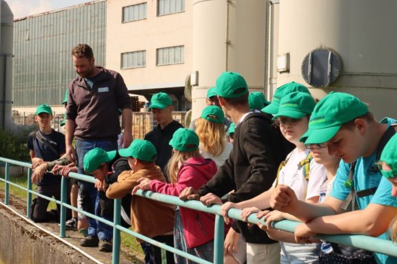 Eine Gruppe von Kindern mit grünen Caps steht an einem Geländer und hört einem Erwachsenen zu.