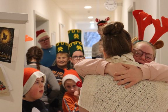 Gruppe von Personen in festlicher Kleidung und Weihnachtsmützen, die sich in einem Flur umarmen.