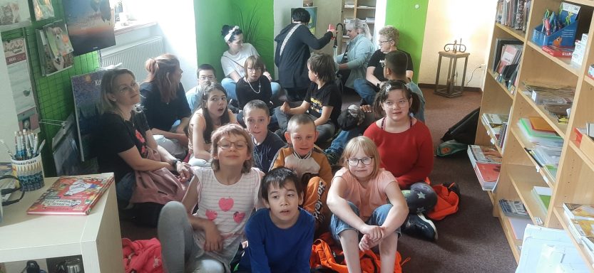 Gruppe von Kindern sitzt in einem Raum, umgeben von Regalen mit Büchern.