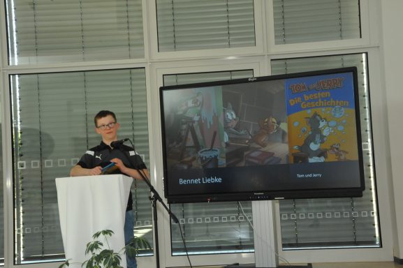 Junger Mann steht am Rednerpult neben einem Bildschirm mit Präsentationsinhalten.