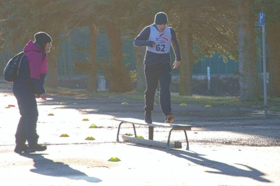 Ein Sportler springt über eine Bank, während eine Zuschauerin zuschaut.