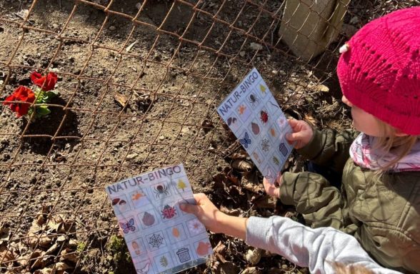 Kinder halten Blätter mit Bilder von Pflanzen vor einem Blumenbeet.