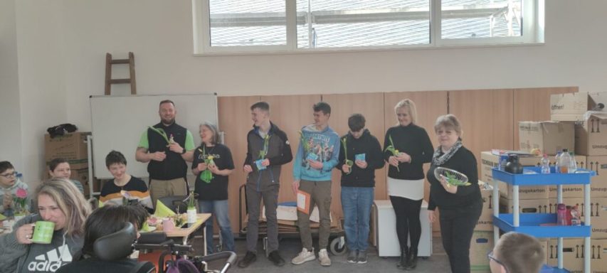 Gruppe von Personen steht in einem Raum, einige halten Tassen, andere sitzen.