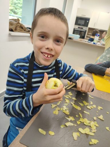 Ein Junge in blauer Kleidung schält einen Apfel und lächelt dabei. Äpfelstücke liegen verstreut.