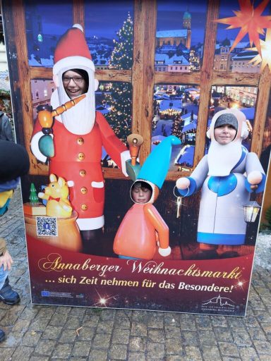 Drei Personen in weihnachtlichen Kostümen stehen vor einem festlich dekorierten Hintergrund.