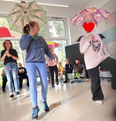 Kinder tanzen mit Regenschirmen in einem hellen Raum. Zuschauer beobachten.