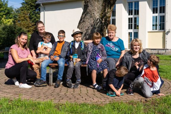Gruppierung von Kindern und Erwachsenen im Freien, lächelnd auf einer Bank sitzend.