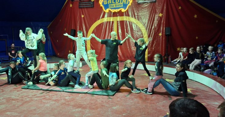 Zirkusaufführung mit Kindern in verschiedenen Kostümen auf der Bühne. Publikum im Hintergrund.