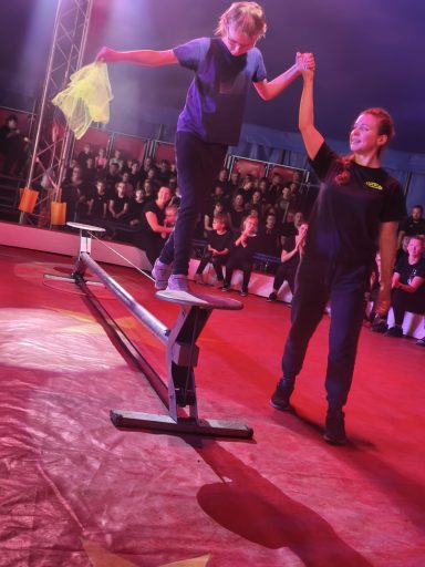 Ein Kind balanciert auf einem schmalen Balken, während eine Trainerin hilft.