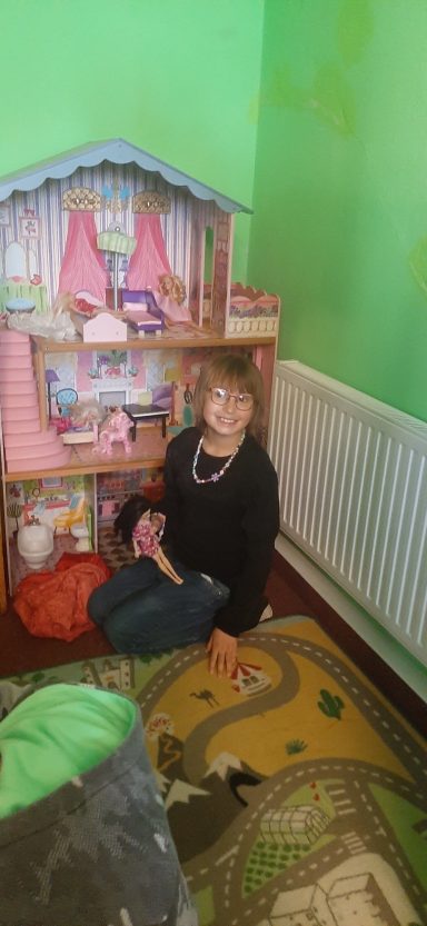 Ein Kind sitzt vor einem Spielzeughaus in einem bunten Raum mit grünem Hintergrund.