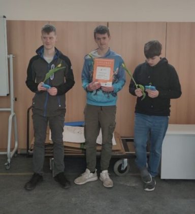 Drei junge Männer halten Urkunden und Pflanzen in einem Raum mit Holzwänden.