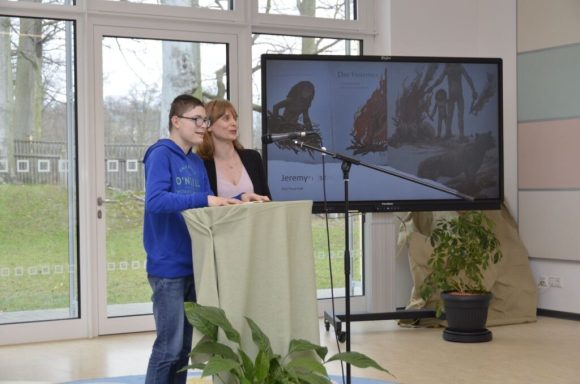 Zwei Personen stehen am Rednerpult, Hintergrund zeigt einen Bildschirm mit einem Bild.