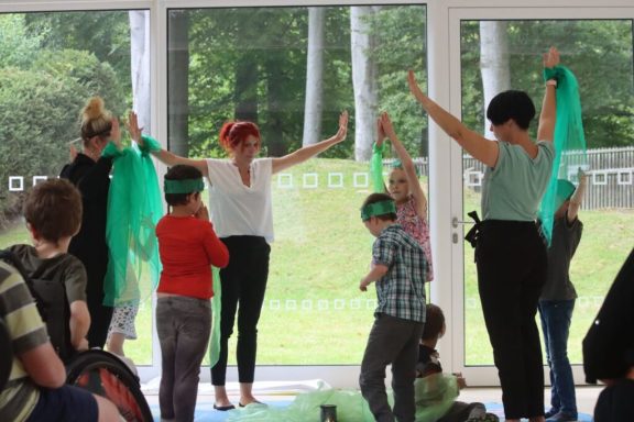 Tanzende Personen mit bunten Tüchern in einem Raum mit großen Fenstern.