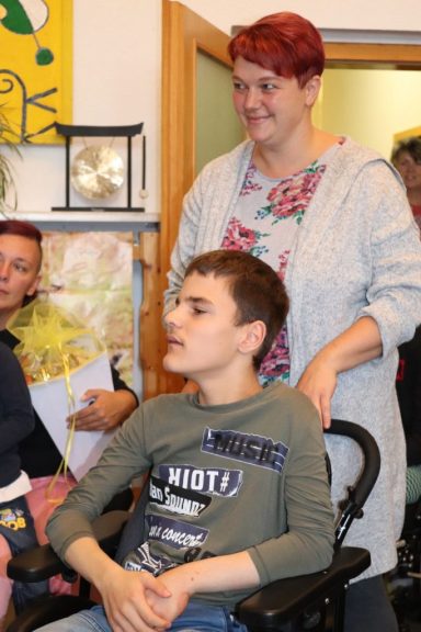 Ein Junge im Rollstuhl sitzt neben einer lächelnden Frau. Im Hintergrund sind Personen sichtbar.