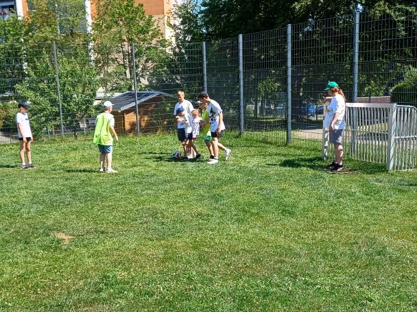 Kinder spielen Fußball auf einem grünen Rasenplatz mit einem Tor im Hintergrund.