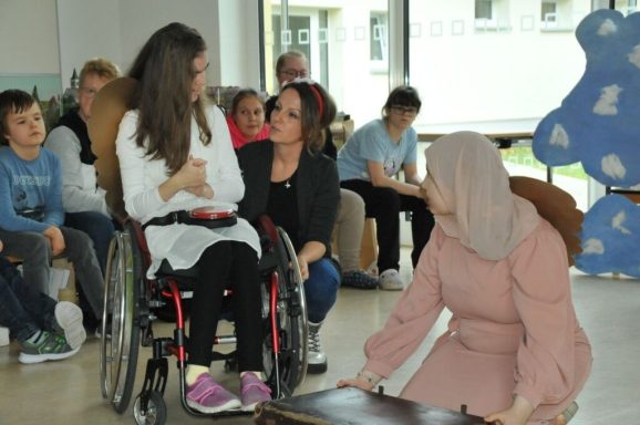 Eine Gruppe von Menschen sitzt zusammen; eine Frau im Rollstuhl spricht mit zwei anderen.