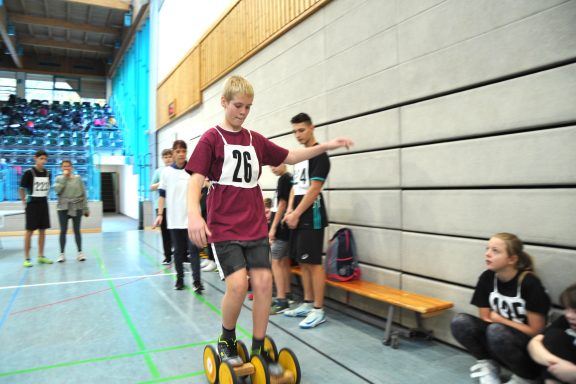 Jugendlicher balanciert auf einem Rollbrett in einer Sporthalle, während andere zuschauen.
