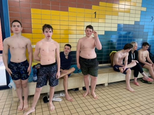 Grupp von Jugendlichen in Badebekleidung in einem Schwimmbad, einige stehen, andere sitzen.