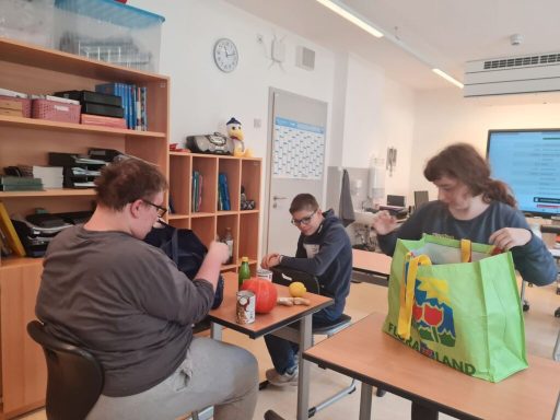 Drei Personen sitzen in einem Klassenraum, arbeiten an Projekten und haben Materialien auf dem Tisch.