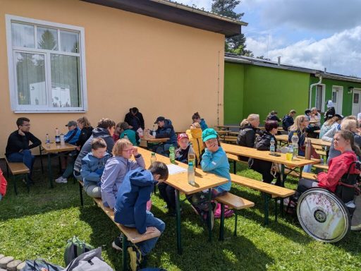 Gruppe von Kindern und Erwachsenen sitzt an Bänken in einem Freiluftbereich.
