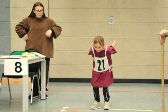 Ein Mädchen mit einer Startnummer springt, während eine Erwachsene zuschaut.