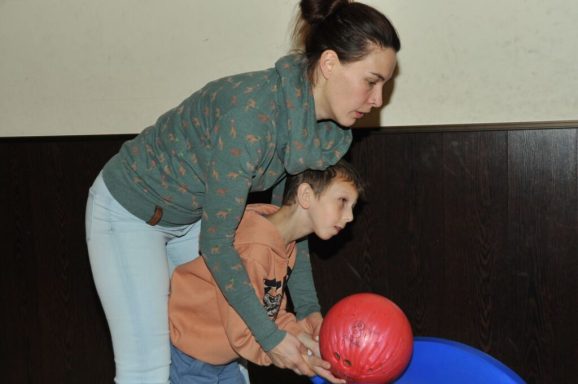 Eine Frau hilft einem Kind beim Bowling mit einer roten Bowlingkugel.