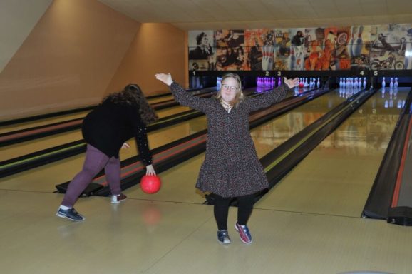 Ein Kind mit braunen Haaren freut sich, während eine Person mit rotem Bowlingball wirft.